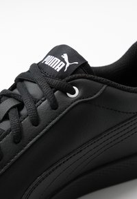 Zapatillas de cuero negro con parte superior texturizada, que presentan cordones negros, una etiqueta de lengüeta blanca con la marca y detalles de costura distintivos.