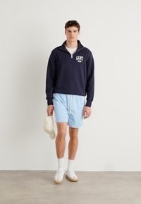 Mørkeblå hettegenser med halv glidelås og logo, lyseblå shorts med snor, hvite sneakers, holder en lys beige caps, står på en nøytral gulv.