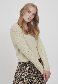 Cremefarvet strikket sweater med v-hals og åben arbejdning på ærmerne, parret med en sort nederdel med blomsterprint.