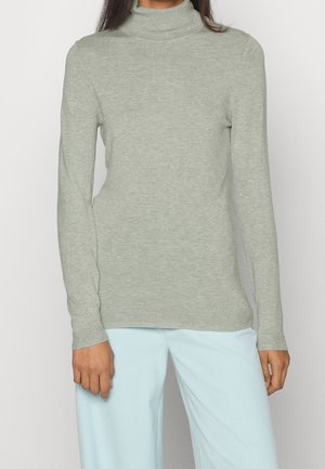 Frau trägt einen hellgrauen Rollkragenpullover und hellblaue Hosen, steht vor einem schlichten weißen Hintergrund.