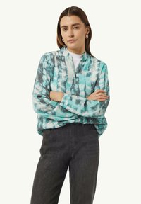 Reißverschluss-Sweatshirt im türkis- und grau-gefärbten Batikmuster. Lange Ärmel mit Bündchen, über einem weißen T-Shirt getragen, kombiniert mit dunklen Jeans.