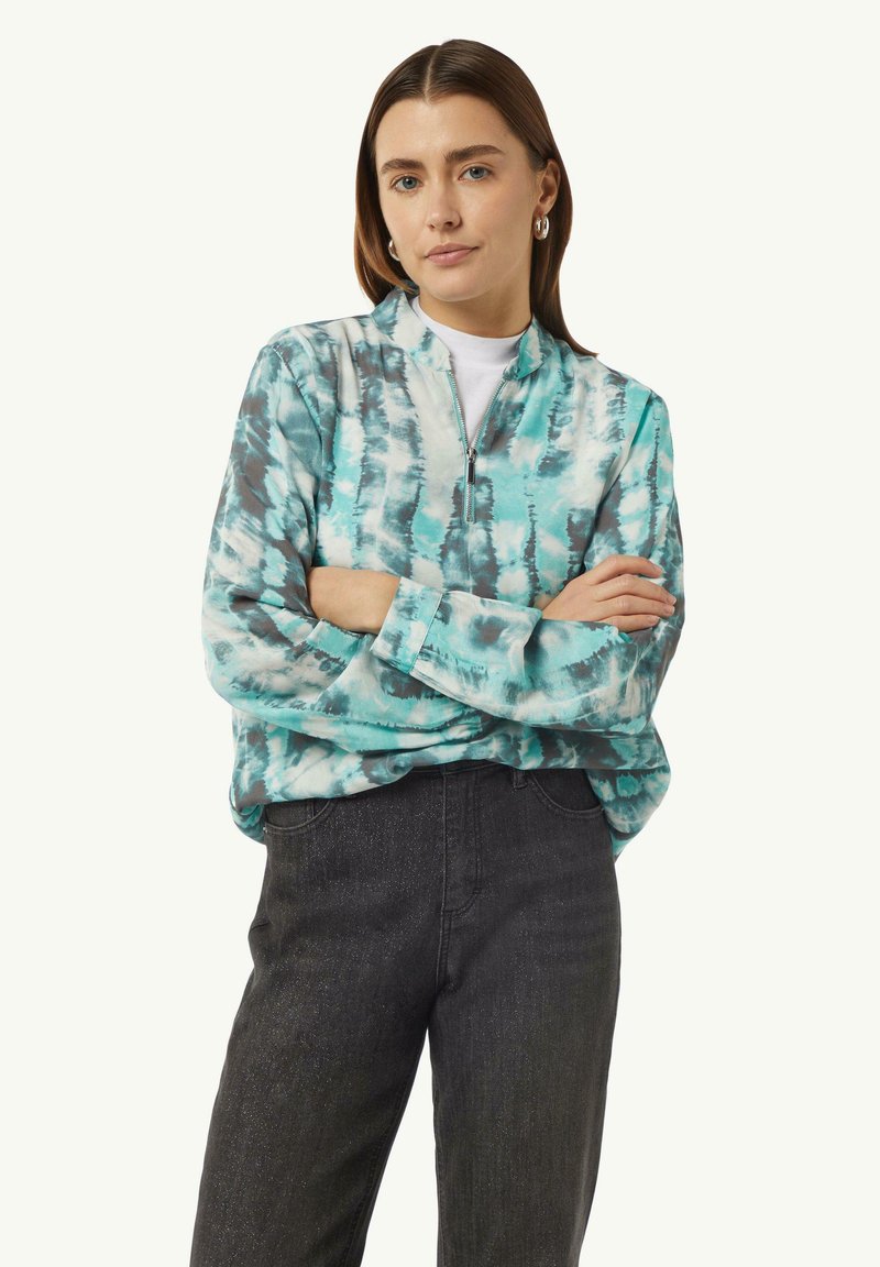 Reißverschluss-Sweatshirt im türkis- und grau-gefärbten Batikmuster. Lange Ärmel mit Bündchen, über einem weißen T-Shirt getragen, kombiniert mit dunklen Jeans.