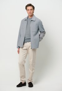 Giacca in misto lana grigio chiaro con colletto a punta, taschino sul petto, abbinata a un maglione grigio e pantaloni beige. Mocassini marroni completano il look.