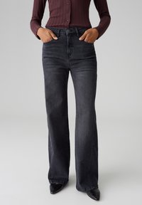 Jean noir à taille haute et évasés avec une texture en denim lisse. Comprend des poches avant et une fermeture à bouton. Porté avec un top ajusté bordeaux.