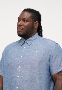 Camisa de chambray azul de manga corta con cuello abotonado, textura sutil, botones blancos y un pequeño logo en el pecho.