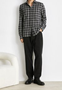 Camisa de cuadros en negro y gris, de corte relajado, de mangas largas, combinada con pantalones negros y botas negras, sobre un fondo neutral.