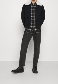 Marine bommerjack met een wit bontkraag, gedragen over een zwart-groen geruite blouse, gecombineerd met zwarte jeans en enkellaarsjes.