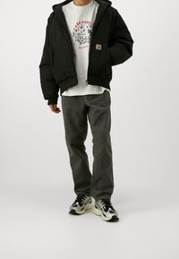 Zwarte gewatteerde jas met capuchon, ritssluiting, vooraan een logo-patch, witte grafische t-shirt eronder, grijze broek en zwart-witte sneakers.