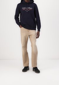 Felpa con cappuccio blu navy con logo bianco e rosso, jeans beige chiaro e sneakers nere con suola testurizzata. Il modello è in piedi contro uno sfondo semplice.