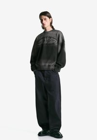 Jongere met schouderlang haar, gekleed in een zwart oversized sweatshirt, brede zwarte broek en zwart glanzende schoenen tegen een witte achtergrond.