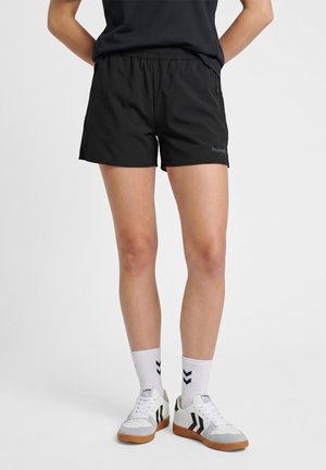 Persoon draagt zwarte sportshorts, witte sokken met zwarte chevron-logo's en witte sneakers met zwarte accenten op een effen achtergrond.