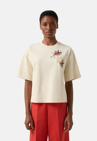 Krémové tričko s krátkými rukávy s kv�ětinovou výšivkou v červené a zelené barvě, s béžovým písmenem "B" na hrudi. Hladká látka.