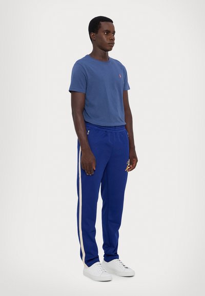 Uomo in piedi con una maglietta blu, pantaloni da jogging blu royal con strisce beige ai lati e sneakers bianche su uno sfondo bianco puro.