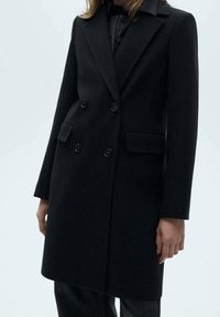 Manteau noir à double boutonnage avec col à revers cranté, deux poches avant et fermetures à boutons, confectionné dans un tissu lisse ressemblant à de la laine.