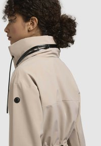 Giacca impermeabile beige con colletto alto, dettagli in hardware neri, coulisse regolabile e texture liscia, con logo sulla manica.
