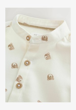 Camisa de botón de color crema claro con pequeños patrones de osos marrones, con un cuello redondeado y dos botones. Fabricada en una tela suave.