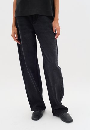 Personne portant un jean noir large et des chaussures noires à bout carré, debout devant un fond clair uni.