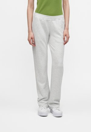 Personne portant un pantalon de survêtement gris clair, des baskets blanches et un haut vert, debout devant un fond blanc uni.