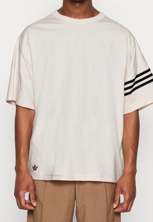 T-shirt oversize beige clair avec un col rond et un design à trois rayures noires sur la manche gauche. Logo minimaliste à l'ourlet.