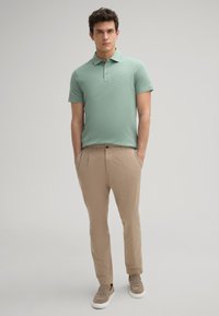 Lichtgroen poloshirt met een kraag en een knoopsluiting van drie knopen, gecombineerd met beige broek. Model draagt textuurbruin instapchoenen.