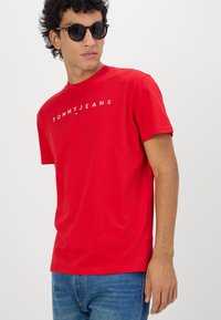 T-shirt en coton rouge à manches courtes, avec le texte blanc "TOMMYJEANS" sur la poitrine. Porté avec un jean en denim bleu et des lunettes de soleil noires.