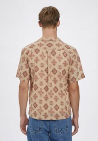 Koszula z krótkim rękawem w kolorze beżowym z geometrycznym wzorem w odcieniach czerwieni i brązu. Posiada prosty kołnierzyk i luźny krój.
