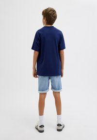 T-shirt in cotone blu navy con maniche corte, abbinato a pantaloni corti in denim chiaro con risvolti. Il modello è in piedi con le spalle rivolte verso la fotocamera.