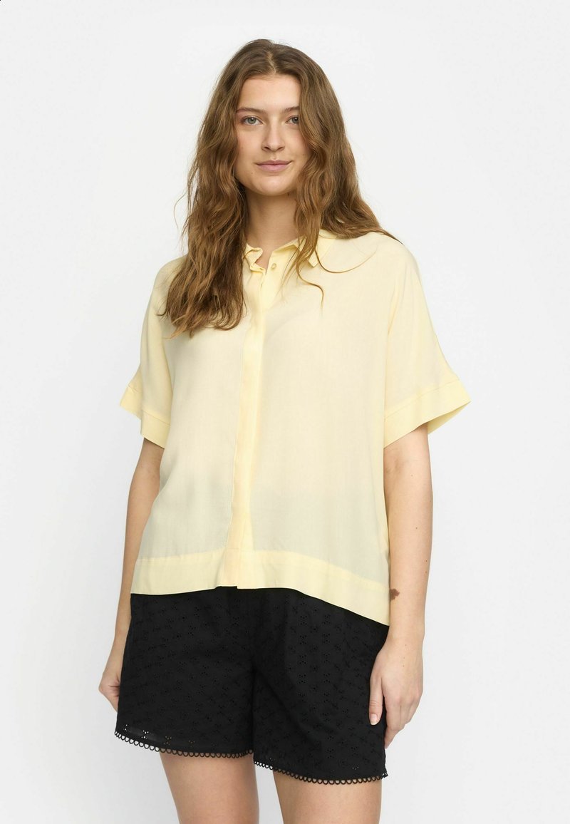 Light yellow short-sleeve blouse with a button front and collar, made from a sheer fabric. Paired with black embroidered shorts.