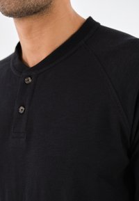 Schwarzes Baumwoll-Henley-Shirt mit einem gerippten Halsauschnitt, einer Zwei-Knopf-Leiste und Raglanärmeln, das eine glatte, weiche Textur aufweist.