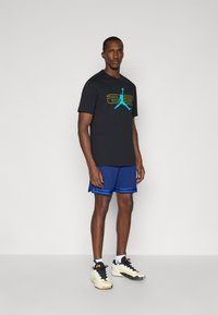 Czarny t-shirt z niebieskim i żółtym wzorem graficznym, w połączeniu z niebieskimi szortami sportowymi z paskowanymi akcentami. Białe sneakersy.