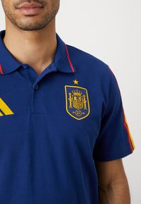 Zila polo krekla ar sarkaniem un dzelteniem akcentiem, uz krūts ar dzeltenu Spānijas nacionālo emblēmu. Izgatavots no teksturēta auduma.