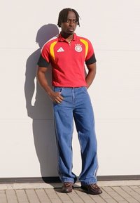 Red and black short-sleeve polo shirt with yellow accents, paired with loose blue jeans and brown shoes. Standing against a light background.