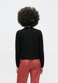 Inimene lokkis afro-soenguga, kandmas musta tekstuuriga pikkade varrukatega pluusi ja punaste mustriliste pükste, selg kaamerale.