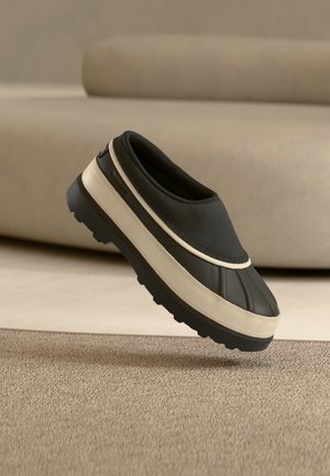 Sort og beige slip-on sko med tyk sål svæver over tæppet, beige sofa uskarp i baggrunden.