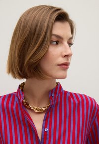 Vrouw met kort lichtbruin haar, gekleed in een rood en paars gestreept shirt en een stevige gouden ketting, kijkend naar rechts.