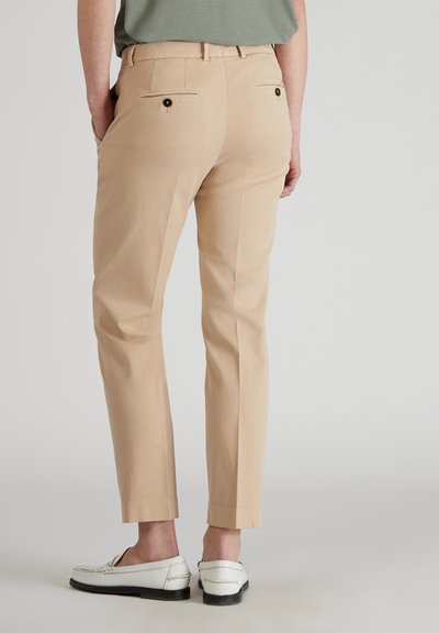Beige broeken met een rechte snit, voorzien van twee achterzakken met knopen, en gecombineerd met witte loafers. De stof lijkt soepel en lichtgewicht te zijn.
