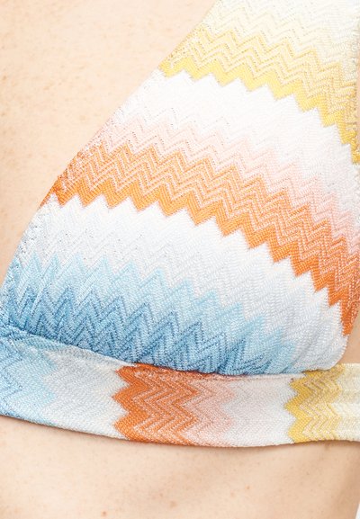 Gros plan sur un haut de bikini en tissu à motif zigzag multicolore dans des nuances de bleu, orange, jaune et blanc sur une peau claire.