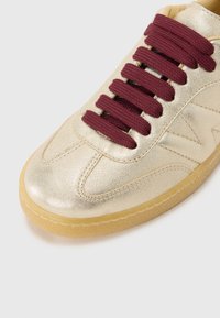 Kultaiset metallin väriset lenkkarit maroonin värisillä nauhoilla, kumipohjalla ja ompeluyksityiskohdilla. Sujuva pinta, pyöreä kärki ja matala muotoilu.