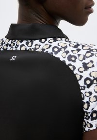 Κοντινό πλάνο ενός ατόμου που φοράει μαύρο πουκάμισο με λεοπάρ σχέδιο, μαύρο γιακά και μικρό λευκό λογότυπο καρδιάς στην πλάτη.