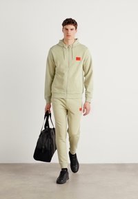 Light green zip-up hoodie and matching joggers with red logo patches. Black sneakers and a black bag are also included in the outfit.
