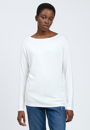 Top blanco de manga larga con escote barco y textura suave, combinado con jeans de mezclilla azul. El top tiene un ajuste holgado y un detalle mínimo.