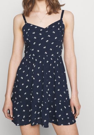 Mujer con vestido sin mangas azul marino con pequeños estampados florales blancos y un dobladillo con volantes, de pie frente a un fondo liso.