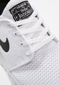 Bílé tenisky Nike z prodyšného materiálu, s černým logem Swoosh, bílými tkaničkami a černou etiketou na jazyku. Texturovaný povrch.