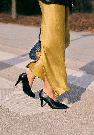 Henkilö kävelee jalkakäytävällä mustissa korkeakorkoisissa kengissä ja laskevassa keltaisessa hameessa pitäen pientä kudottua sinistä käsilaukkua.