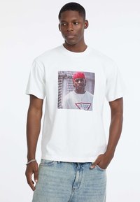 Biała bawełniana koszulka z krótkim rękawem, z grafiką na przodzie przedstawiającą osobę w czerwonej czapce. Na dole noszone są zniszczone dżinsowe szorty.