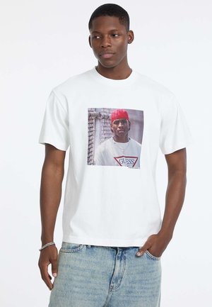 Weißes Baumwoll-T-Shirt mit kurzen Ärmeln, mit einem Frontdruck von einer Person, die eine rote Mütze trägt. Darunter wird eine zerrissene Jeansshorts getragen.