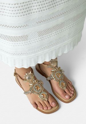 Voeten met beige sandalen versierd met grote edelstenen en schelpen, gecombineerd met een witte, gestructureerde rok.