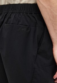 Gros plan d'une personne portant un short à taille élastique noire avec des poches latérales et arrière, la main reposant près de la cuisse.