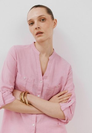 Camicia di lino rosa chiaro con colletto alla cinese, due tasche sul petto e polsini abbottonati, indossata da un modello con le braccia incrociate.