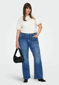 Žena v bílém topu s krátkým rukávem a krajkovým lemem, modrých vysokých rozšířených džínách, černých lodičkách a držící černou kabelku se cvočky.